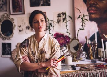 Beautiful young woman artist looking away thoughtfully with her arms crossed and holding some paintbrushes in her creative studio wearing an old-fashioned artist's smock which is covered with paint marks