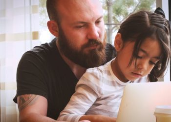 A young caucasian man in his 20s spending quality bonding time with a little 5 year old hispanic girl with long black hair and brown eyes as he teaches her how to use a laptop computer in a well lit cheerful room