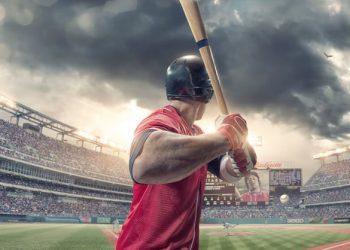A close up rear view of a professional male baseball player wearing a red baseball strip and safety helmet, holding baseball bat just about to strike incoming baseball. The action occurs during a baseball game in a generic floodlit outdoor stadium full of spectators. Baseball ground, scoreboard etc are all fictional.