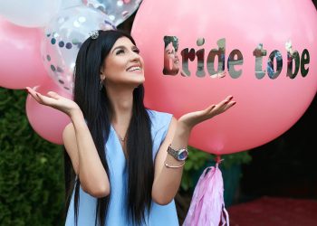 Happy young beautiful brunette bride to be with dark hair and silver crown smiling and gesturing spread out her hands in joy on bachelorette pink party balloon background.