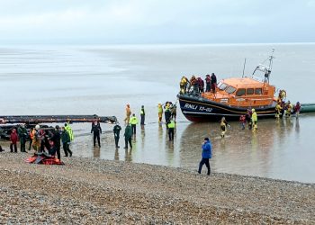 A lifeboat crew assist migrants from the English Channel in Dungeness, Kent.  November 20, 2021.  See SWNS story SWOCmigrants.  Hundreds of migrants were rescued from the sea by RNLI lifeguards after crossing the Channel in small boats.  Adults and young children wrapped in blankets were seen being led up the beach in Dungeness, Kent on Saturday (20/11).  They were helped by lifeguards from RNLI Dungeness after attempting to seek asylum before the winter weather makes the journey too difficult.  At least four dinghies packed with dozens of people reportedly reached the shore, as well as a lifeboat bringing in more from out at sea.  Others arrived at Folkestone and Dover harbours, brought in by authorities.  Border Force agents and police were on the scene as well as three coaches to pick the migrants up and take them to be processed.